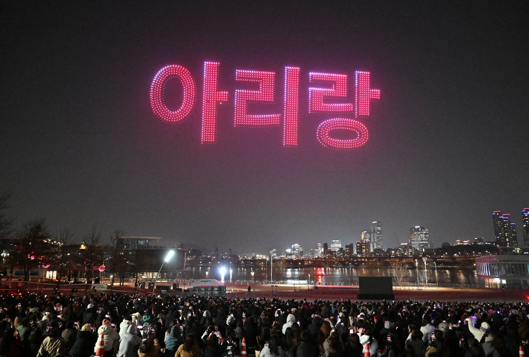 The word Arirang in red Hangul letters floats in red over the night sky over a city