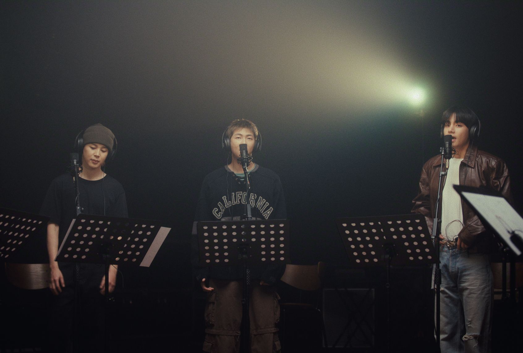 Three men with headphones stand in front of music stand as the stand in a black studio recording vocals