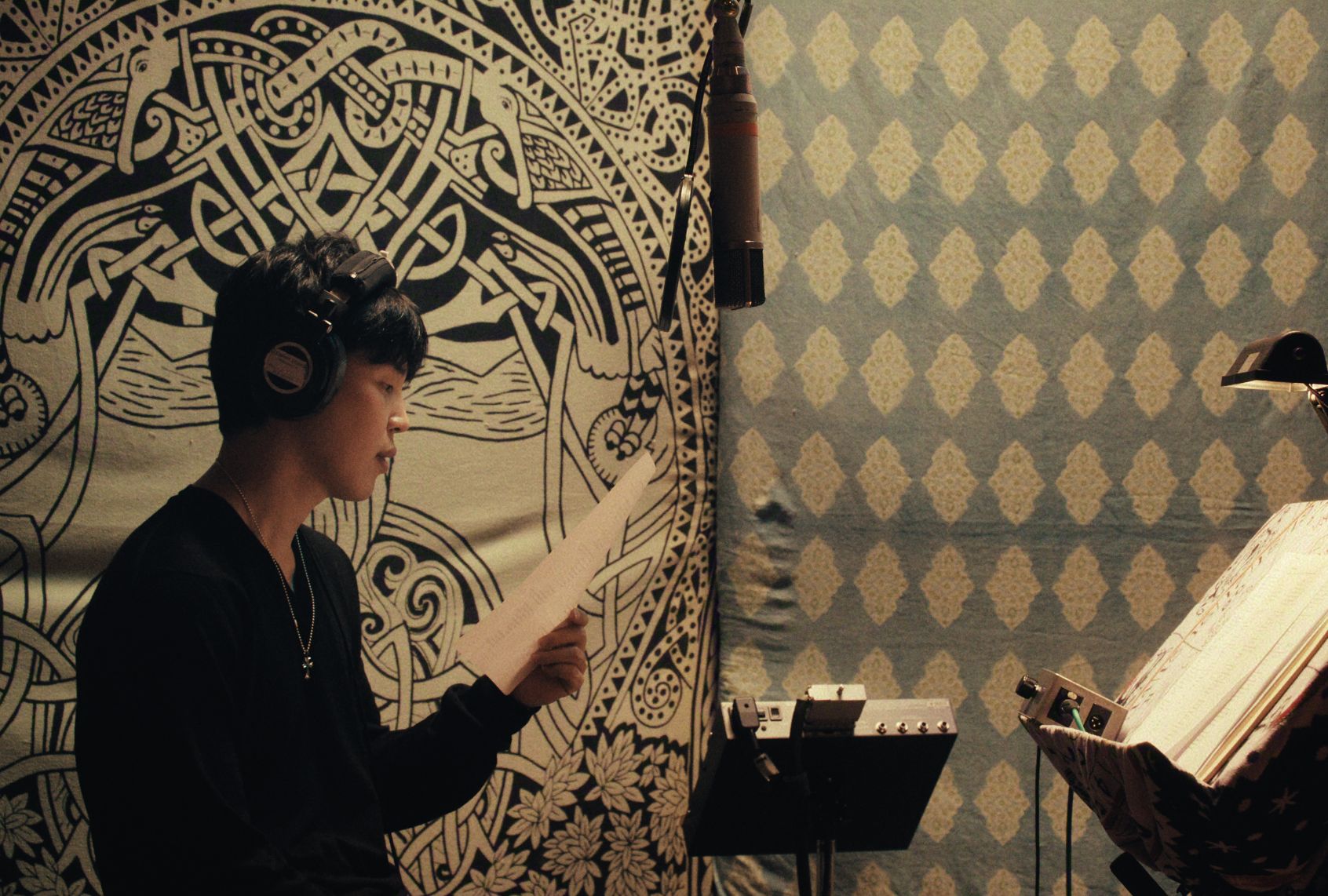 A man in a black sweater and black headphones on reads from a sheet of paper in his hand. He appears to be a in studio, with a microphone hanging from the ceiling and a patterned wall behind him