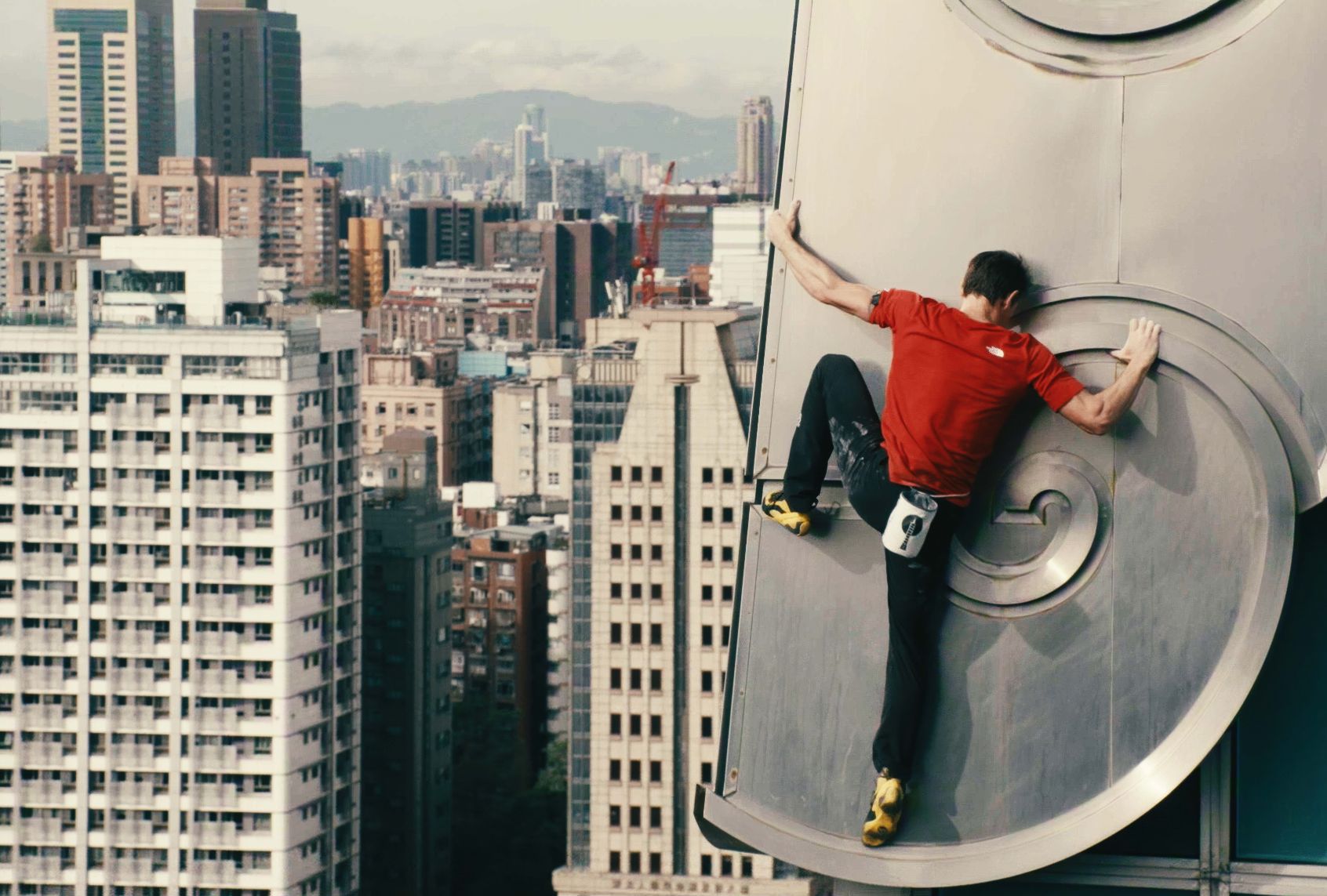A man in a red shirt, dark jeans and yellow shoes has a white chalk bag on his hip. He's holding on for dear life with just his hands and feet on the side of a building. He's very very high up