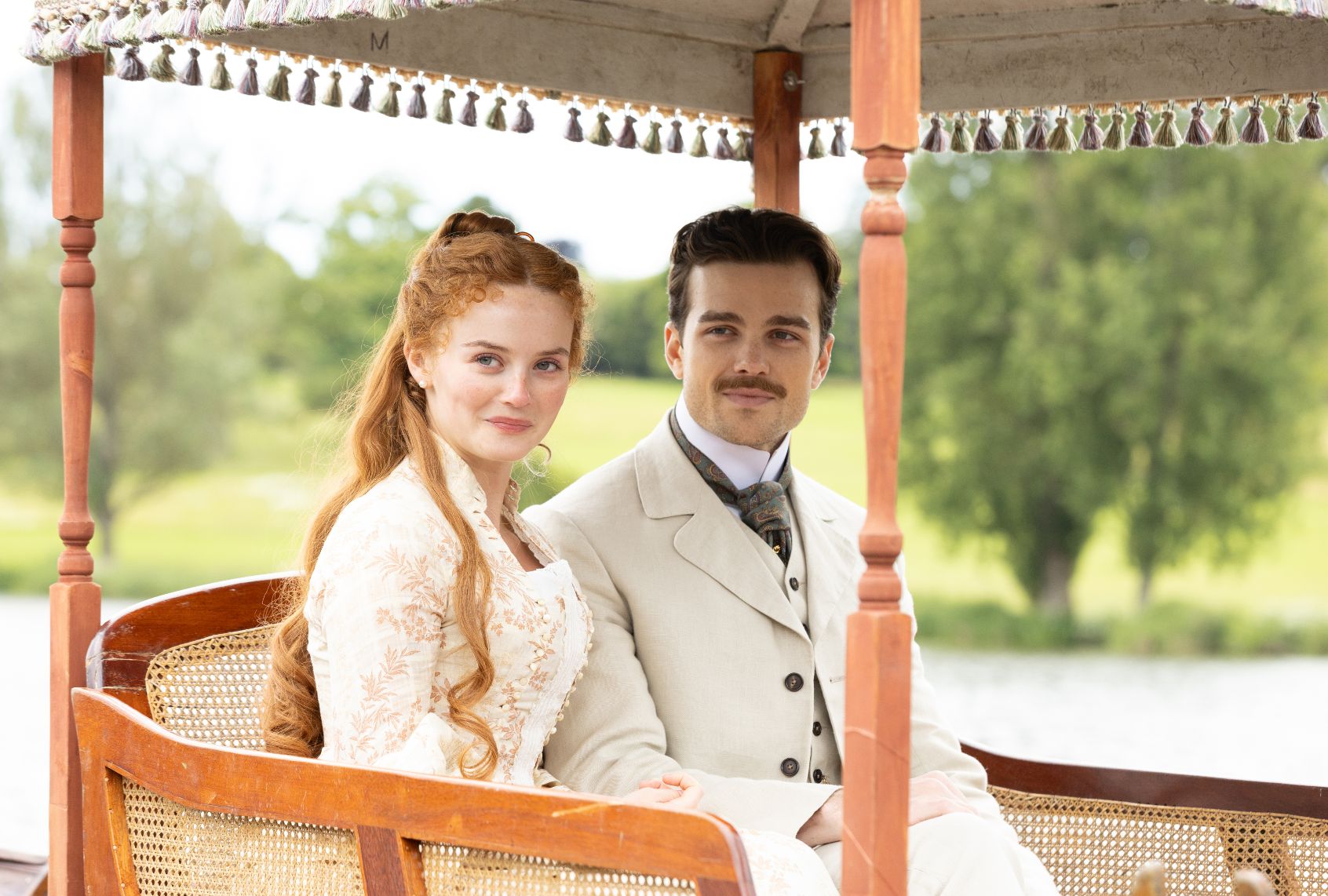 A brunet man with a mustache wearing an off-white linen suit and a redheaded woman with long hair wearing a cream and peach brocade gown sit in what looks to be a wicker surrey (with a fringe on top)