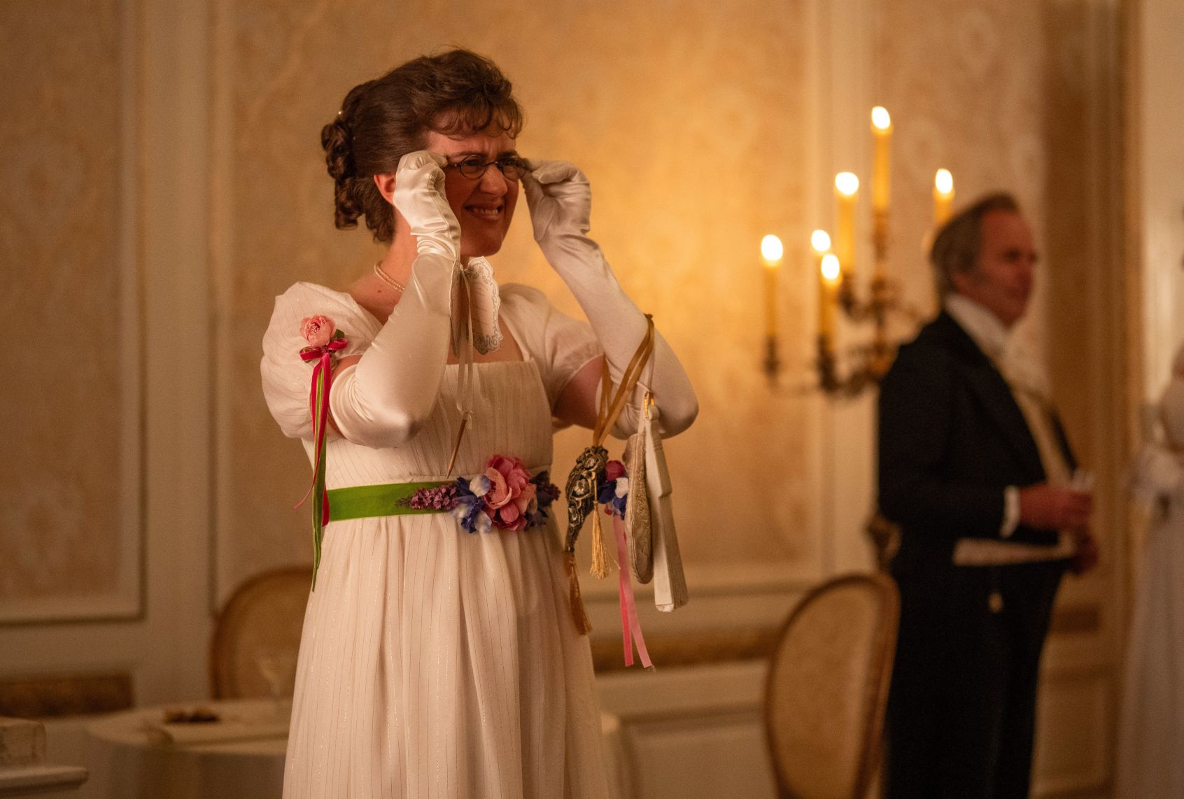 A brunette woman in a high-waisted white gown adorned with ribbons and flowers wears matching elbow-length gloves. She is squinting as she adjusts her wireframe spectacles. She's in a candlelit ballroom with a gentleman in suit in the background