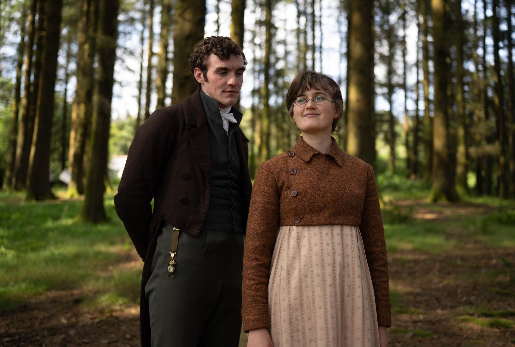 A brunette woman in spectacles, high-waisted jacket and long full skirt smiles as a curly-haired young man in ascot, vest, jacket and breeches gazes at her affectionately. They're standing in a green forest