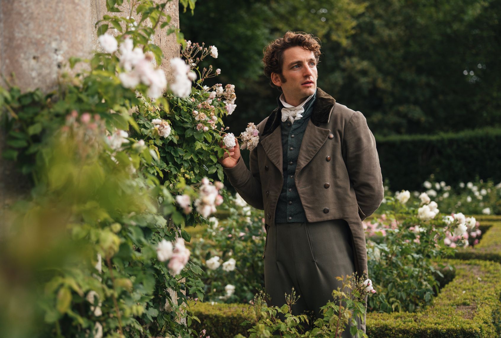 A man in Regency-era ascot, waiscoat, jacket and breeches stands in a flower garden