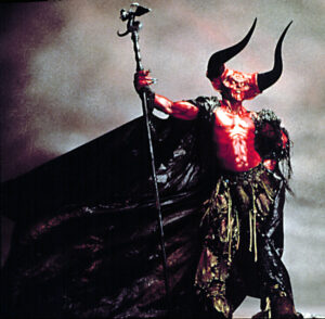 Tim Curry in full red, horned makeup and prosthetics as the Lord of Darkness in "Legend," holding a staff aloft, with a large black cape draped behind him.