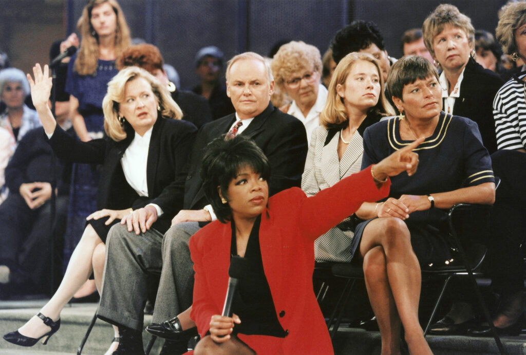 Oprah Winfrey, wearing a red blazer and holding a microphone, kneels in front of rows of seated audience members and points to the side as a woman behind her raises her hand.