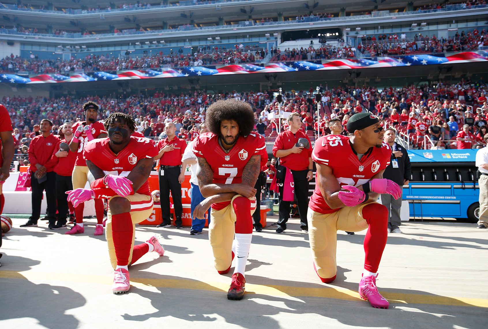 Three San Francisco 49ers players in red jerseys kneel on the sideline during the national anthem as teammates and fans stand behind them.
