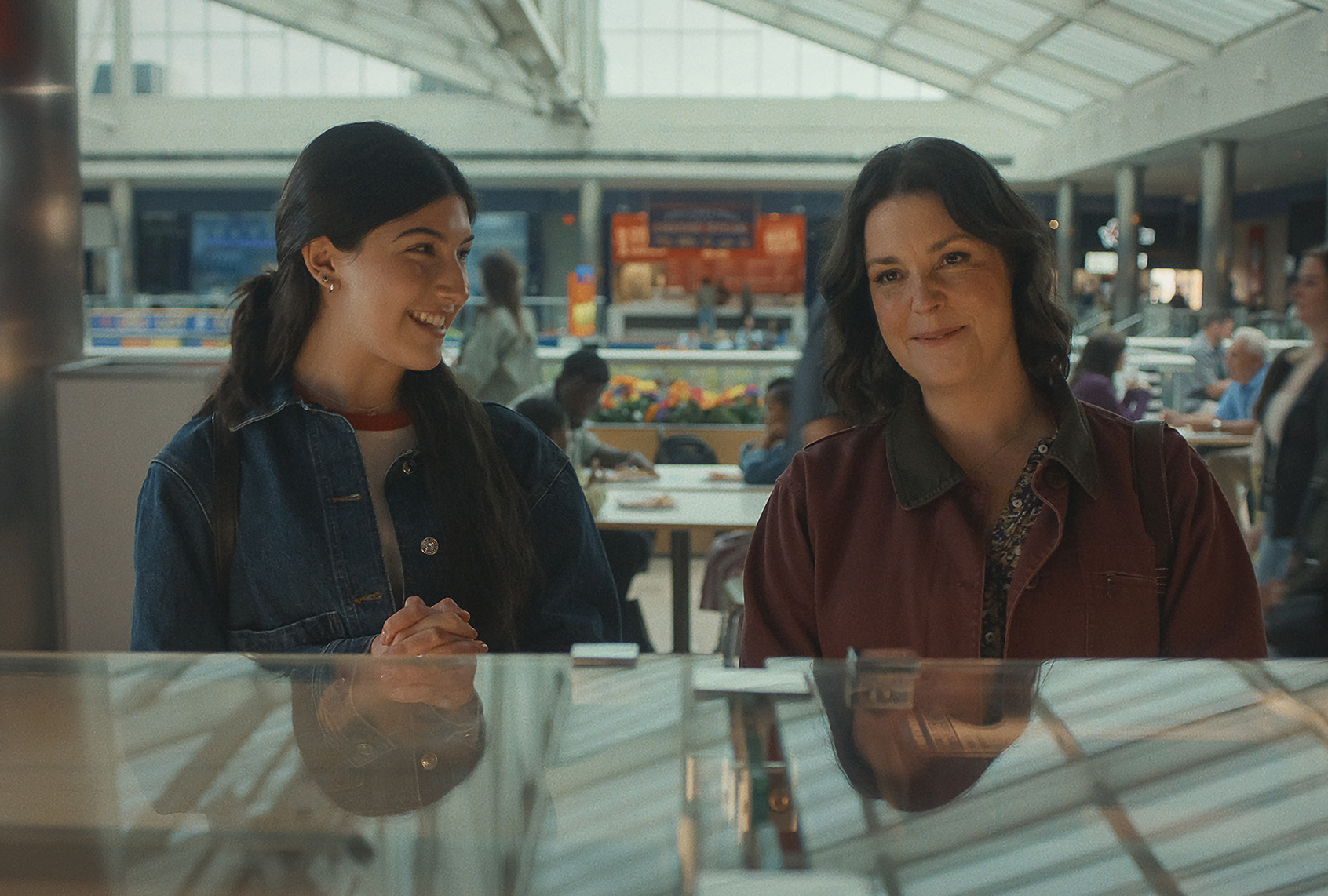 Two women stand together at a jewelry counter in a mall food court, one in a denim jacket smiling and the other in a maroon jacket looking on, in a scene from "Don't Say Good Luck."