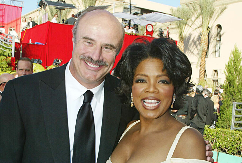 Phil McGraw, in a black suit and black tie, stands with Oprah Winfrey, in a yellow dress and drop earrings, as both smile on a red carpet with red staging, palm trees and photographers behind them.