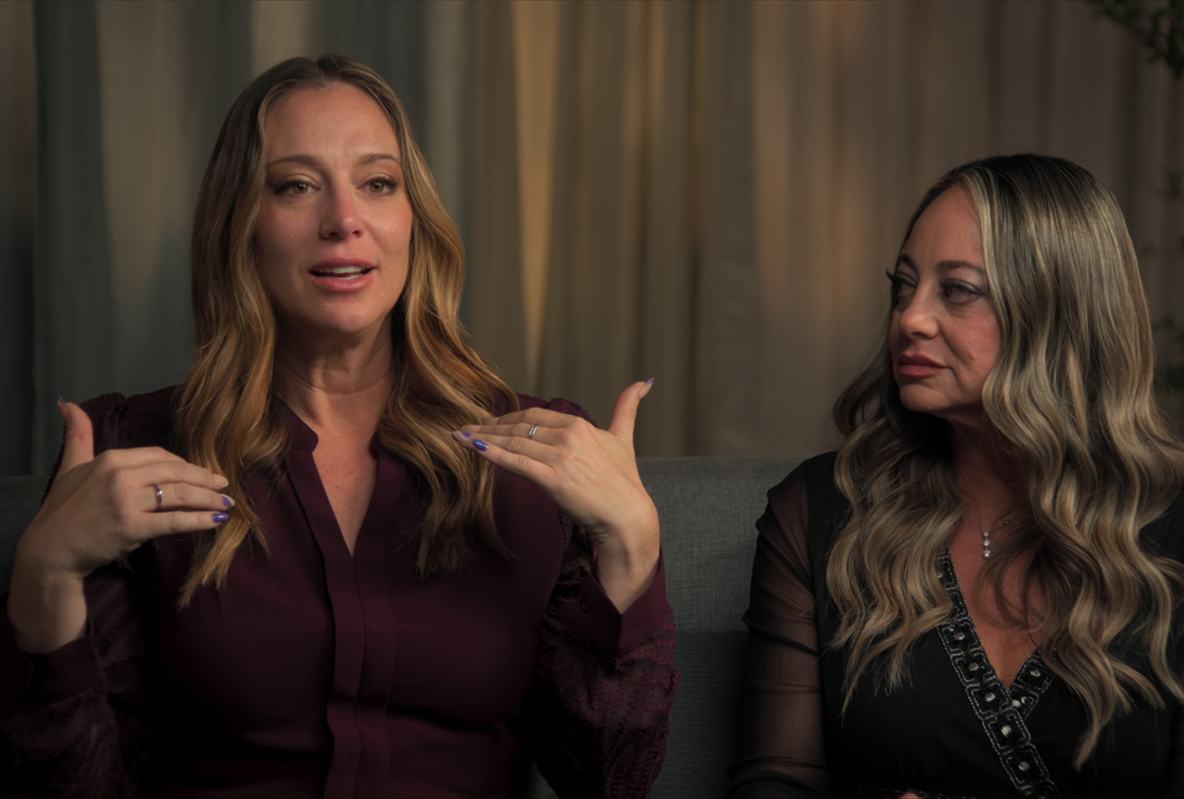 Two women sit side by side on a gray couch in front of beige curtains. The woman on the left, wearing a burgundy blouse, speaks and gestures toward herself with both hands. The woman on the right, wearing a black top with beaded trim, turns to look at her.