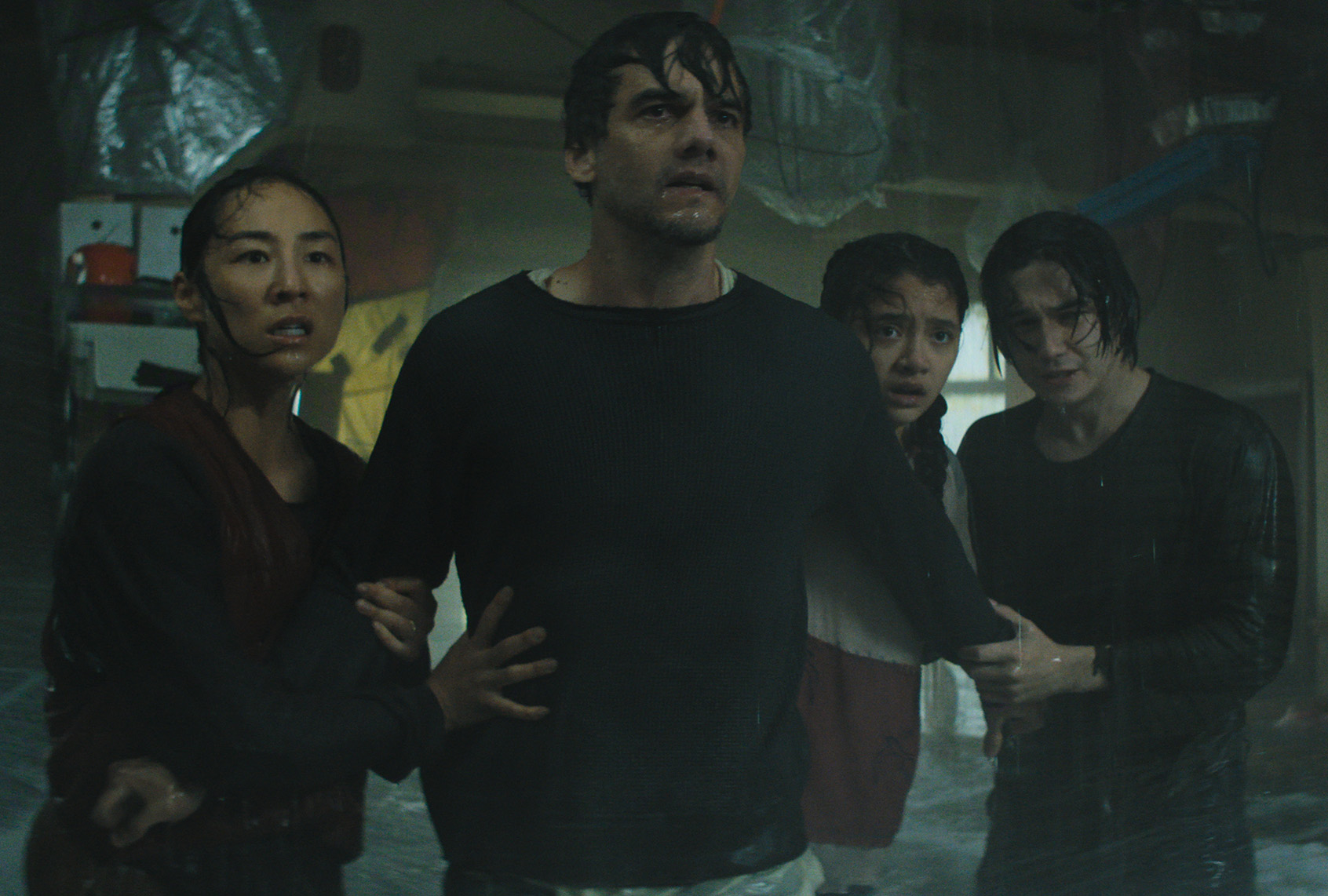 A family of four stands close together in a dark, water-damaged room, their clothes soaked and expressions fearful, in a scene from 