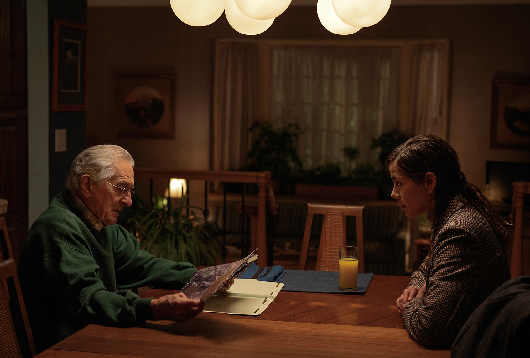 A dimly lit dining room scene from "The Whisper Man" showing Robert De Niro and Michelle Monaghan seated at a table, examining a file folder as she looks on with a serious expression.